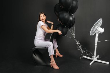 Portrait of Young brunette sitting and holding bunch of black balloons when a fan blowing on it, isolated on black background.