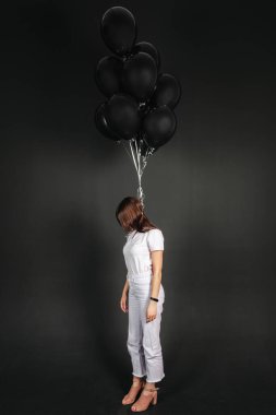 Portrait of a young brunette standing with her face covered by her hair and lifted black balloons tied to her neck, isolated on black background.