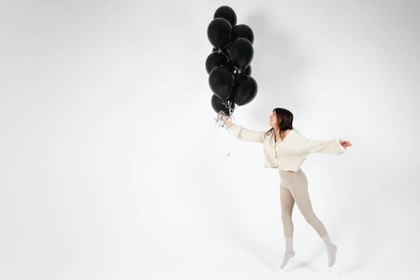Brunette Girl in light clothes jumping with Bunch of black balloons. Isolated on white background.