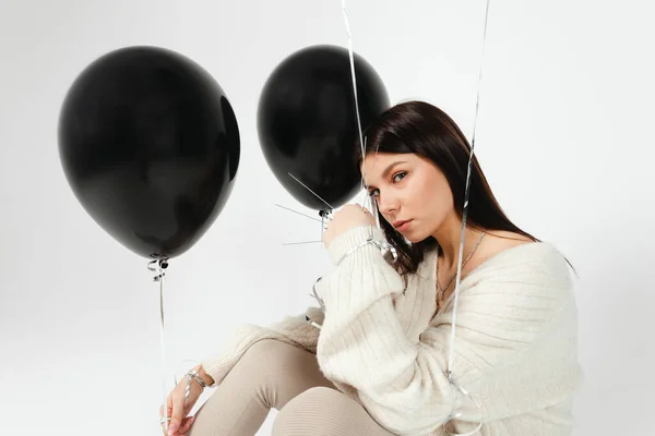 Portrait of Young brunette holding black balloons and full hand of needles ready to pop them, isolated on white background. letting go of negative.