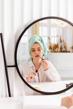 A pretty girl is sitting in front of a mirror with a towel wrapped around her head, doing her makeup.