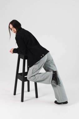 Portrait of a beautiful brunette girl leaned on a bar chair, dressed in a black jacket, wide jeans. Isolated on white.