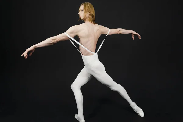 Young athletic professional ballet dancer with a bare torso and white dance tights is in perfect shape and posing with bright yellow slippers in his hands over a black background.