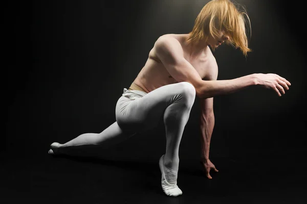 Young athletic professional ballet dancer with a bare torso and white dance tights is in perfect shape and posing with bright yellow slippers in his hands over a black background.