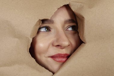 Close up: A girl's face is peeking out from a cutout in a large sheet of kraft paper.