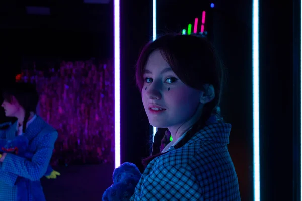 A cute girl with drawn tears, dressed in a blue plaid jacket , holds soft toy and looks at the camera in a neon-lit room.
