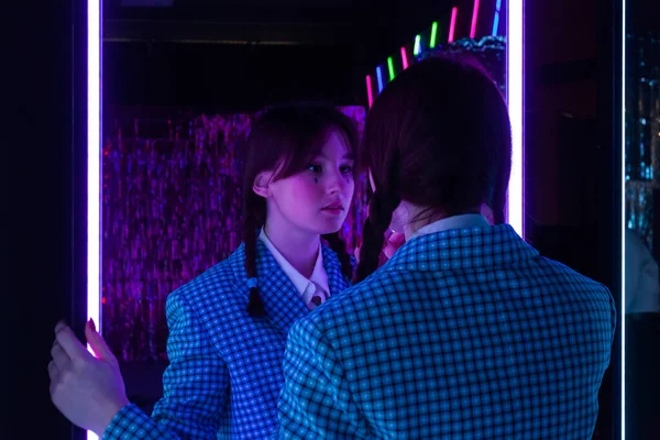 A cute girl with drawn tears, dressed in a blue plaid jacket and skirt with knee high socks, looks mysteriously at herself in the mirror in a neon-lit room.