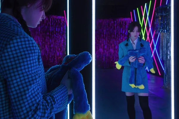 A cute girl with drawn tears, dressed in a blue plaid jacket and skirt with knee high socks, holds soft toy and looks mysteriously at herself in the mirror in a neon-lit room.
