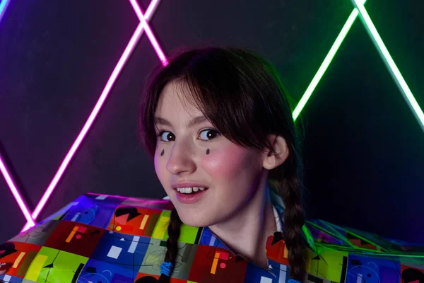 Portrait of a cute smiling girl with drawn tears, dressed in a blue plaid jacket,  peeking out from a cutout in a sheet of glitter wrapping paper in a neon lit room.