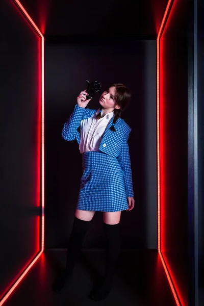 A cute girl with drawn tears, dressed in a blue plaid jacket and skirt with knee high socks, holds black plastic hand in a red neon lit room.