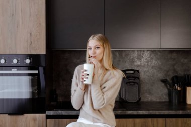 Güzel, ince, sarışın bir kız modern bir mutfakta oturmuş pipetli bir bardaktan soda içiyor..