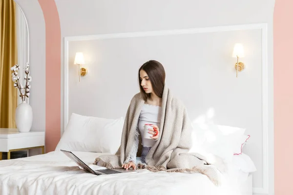 マグカップで暖かい飲み物とノートパソコンで作業している美しい女の子が毛布に包まれたベッドの上に座って — ストック写真