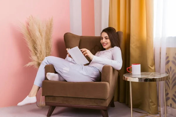 椅子に座り 温かい飲み物を手に本を読む 美しく長髪で幸せな女の子の肖像画 — ストック写真