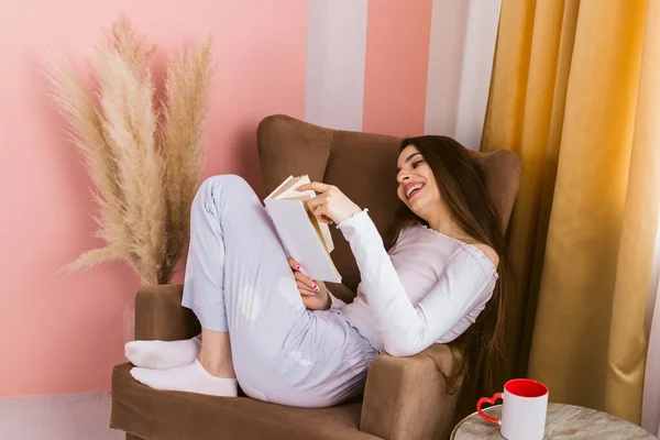 椅子に座り 温かい飲み物を手に本を読む 美しく長髪で幸せな女の子の肖像画 — ストック写真