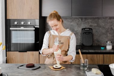 Bej önlüklü genç, gülümseyen bir pasta şefi, modern bir mutfaktaki bir boru torbasından çikolata süngerli pasta tabakasına krema sürerek küçük bir pasta hazırlıyor..