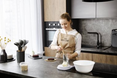 Bej önlüklü genç, gülümseyen bir pasta şefi küçük bir pasta hazırlıyor, modern bir mutfakta çikolata süngerli pasta tabakasına şerbet sürüyor..