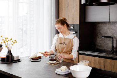 Bej önlüklü genç, gülümseyen bir pasta şefi küçük bir pasta hazırlıyor, modern bir mutfakta çikolata süngerli pasta tabakasına şerbet sürüyor..