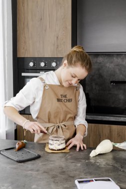 Bej önlüklü genç, gülümseyen bir pasta şefi modern bir mutfakta küçük bir pasta hazırlıyor. Kapat..