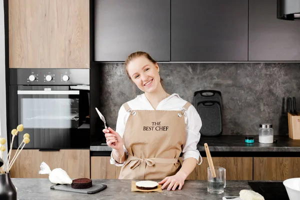 Bej önlüklü genç, gülümseyen bir pasta şefi, modern bir mutfakta küçük bir pasta hazırlıyor..