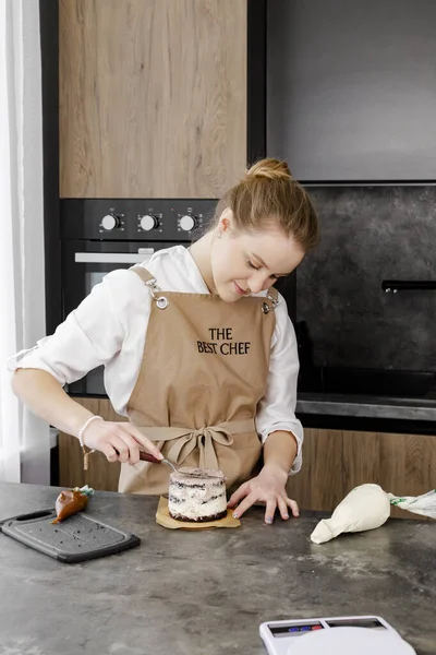 Bej önlüklü genç, gülümseyen bir pasta şefi modern bir mutfakta küçük bir pasta hazırlıyor. Kapat..