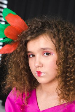 Çekici, kıvırcık saçlı bir kızın portresi, renkli tüylerle süslenmiş, siyah bir odada pembe bir elbise giyiyor..