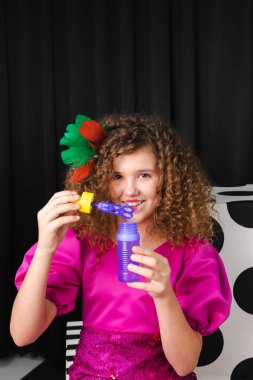 Renkli tüylerle süslenmiş, küçük kıvırcık saçlı bir kızın portresi pembe bir elbise giyer ve siyah bir odada sabun köpüğü üfler..