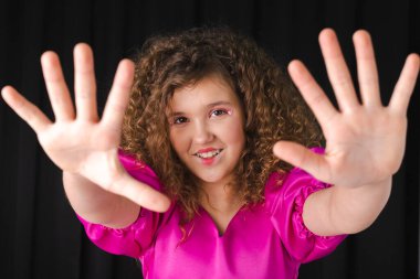 Çekici, kıvırcık saçlı bir kızın portresi, pembe bir elbise giyiyor ve yaratıcı yüz süslemeleri yapıyor, ellerini siyah bir odada öne koyuyor..