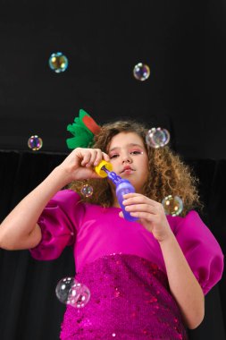 Renkli tüylerle süslenmiş, küçük kıvırcık saçlı bir kızın portresi pembe bir elbise giyer ve siyah arka planda sabun köpüğü sallar. Alt görünüm.