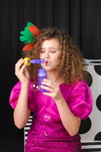 Renkli tüylerle süslenmiş, küçük kıvırcık saçlı bir kızın portresi, pembe bir elbise giyiyor, siyah bir odada sabun köpükleri üflüyor..