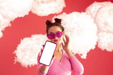 Pembe bluzlu ve kısa etekli çekici, parlak ve mutlu bir kız bulutlu izole bir arka planda boş bir ekran ya da ekran ya da cep telefonu modeli gösterir. Uygulama öneriliyor. Bulut teknolojisi.