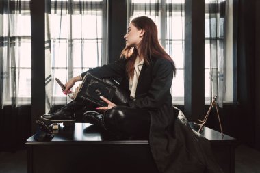 Siyah elbiseli güzel bir kız uzun bir ceket, dar deri tozluklar ve uzun bağcıklı tombul çizmeler giyer. Elinde bir kitapla karanlık bir mahsul odasında oturuyor..