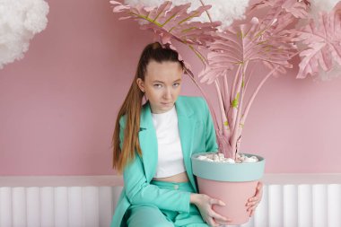 Malakit renginde bir iş elbisesi içindeki güzel bir kız üzerinde pembe bir saksıyla kaplanmış bir vazo içinde yüzen bulutlarla birlikte pastel pembe bir odada..