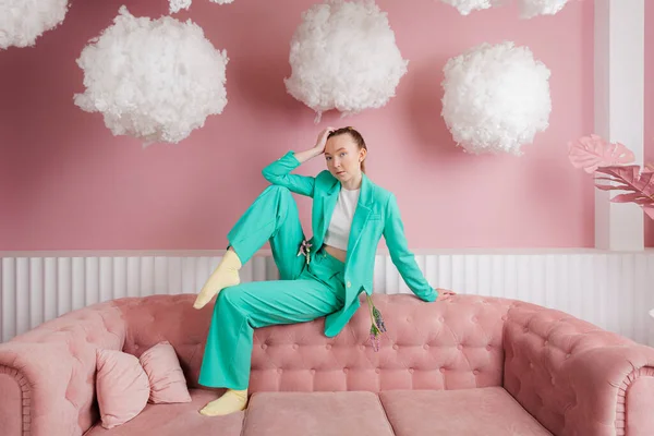 stock image A beautiful girl in a business suit of malachite color sits on a couch or sofa in a pastel pink room with floating clouds.