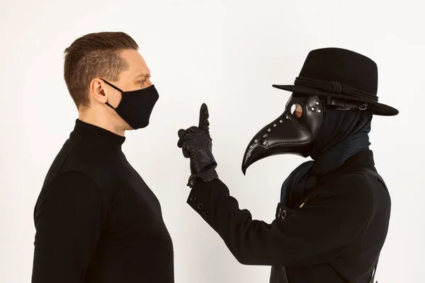 A Plague doctor and a man in a Medical mask looking at each other. a syringe and needle with Medicine or serum, antidote. Isolated on a white background. COVID-19, epidemic and pandemic concept.