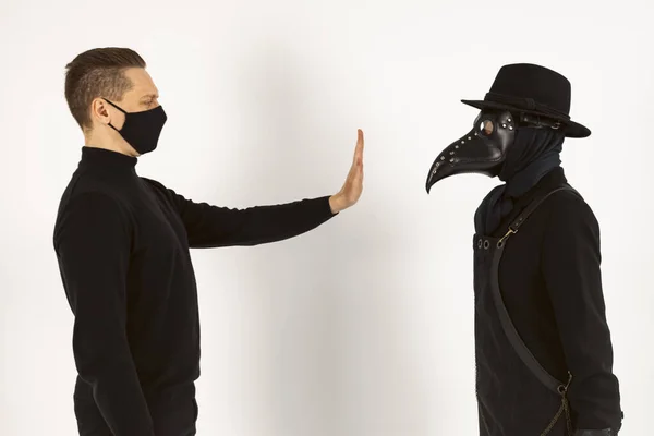 A Plague doctor and a man in a Medical mask gestures not to approach. a syringe and needle with Medicine or serum, antidote. Isolated on a white background. COVID-19, epidemic,pandemic concept.