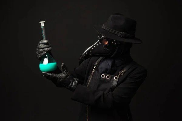 Close-up Portrait of a plague doctor from Medieval era holds a glass vessel containing a potion or serum, antidote. Isolated on a black background. COVID-19, epidemic and pandemic concept.