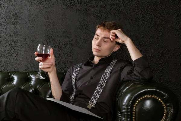 A young man is sitting on an elegant couch in dark room with a glass of wine in his hand and music sheets and contemplating a composition. The process of creation.