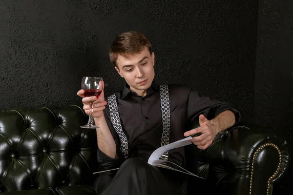 A young man is sitting on an elegant couch in dark room with a glass of wine in his hand and music sheets and contemplating a composition. The process of creation.