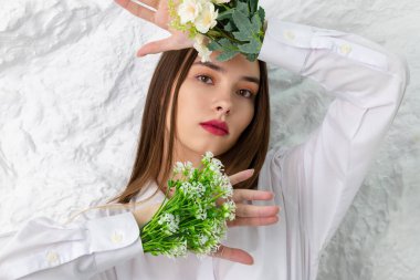 Beyaz gömlekli, elinde çiçeklerle güzel kahverengi gözlü bir kızın portresi. Arka planda beyaz bir duvar var. Güzellik ve Tazelik kavramı.