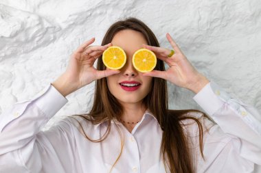 Beyaz gömlekli ve limon dilimleriyle poz veren güzel kahverengi gözlü bir kızın portresi. Arka planda beyaz bir duvar var. Güzellik ve Tazelik kavramı.