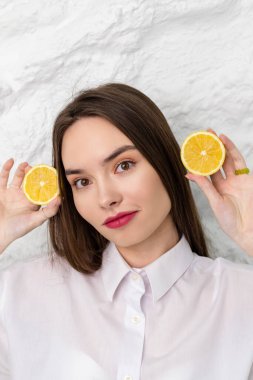 Beyaz gömlekli ve limon dilimleriyle poz veren güzel kahverengi gözlü bir kızın portresi. Arka planda beyaz bir duvar var. Güzellik ve Tazelik kavramı.