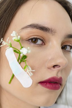 Yakın plan. Yanağına alçı bağlanmış bir çiçekle güzel ve sevimli kahverengi gözlü bir kızın portresi. Arka planda beyaz bir duvar var. Güzellik ve Tazelik kavramı.