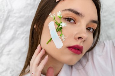 Yakın plan. Yanağına alçı bağlanmış bir çiçekle güzel ve sevimli kahverengi gözlü bir kızın portresi. Arka planda beyaz bir duvar var. Güzellik ve Tazelik kavramı.