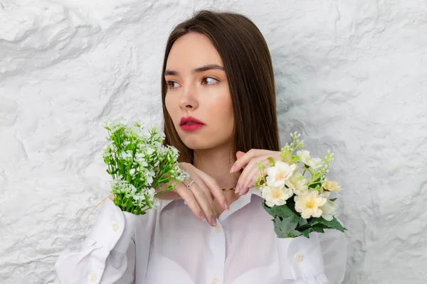 Beyaz gömlekli, elinde çiçeklerle güzel kahverengi gözlü bir kızın portresi. Arka planda beyaz bir duvar var. Güzellik ve Tazelik kavramı.