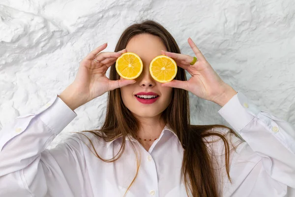 Beyaz gömlekli ve limon dilimleriyle poz veren güzel kahverengi gözlü bir kızın portresi. Arka planda beyaz bir duvar var. Güzellik ve Tazelik kavramı.