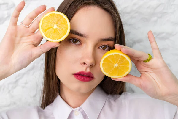 Portrait of a young beautiful brown-eyed girl in a white shirt holding and posing with round slices of lemon. A white wall is in the background. Beauty and Freshness concept.