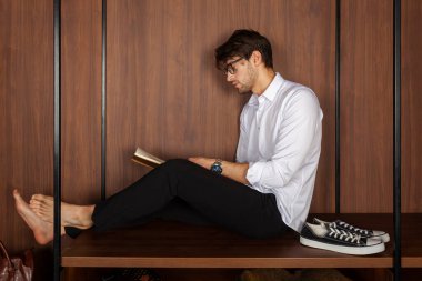 Elbise rafında oturmuş kitap okuyan şık giyinmiş yakışıklı bir genç. yan görünüm.