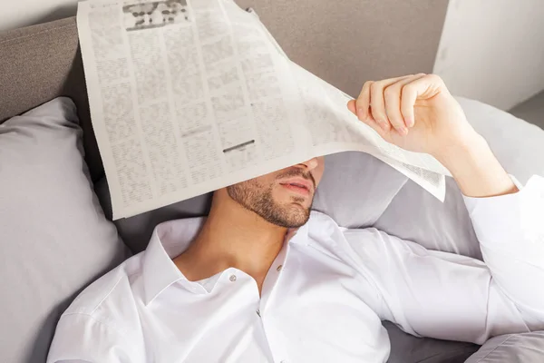 一个穿着白衬衫的英俊年轻人在看报纸时在床上睡着了 满脸都是报纸 — 图库照片