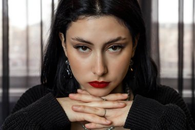 Siyahlı güzel, zarif bir kızın portresi, parmakları çapraz oturur ve çenesi üstlerinde dinlenir ve yüzünde ciddi bir ifade, dikkatlice bakar..