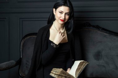 Siyah elbiseli, güzel bir kızın portresi, parlak kırmızı rujlu, elinde bir kitapla koltukta oturuyor ve yüzünde mutlu bir gülümseme ifadesi var, yukarı bakıyor. Karanlık Gotik Vingare Odası.